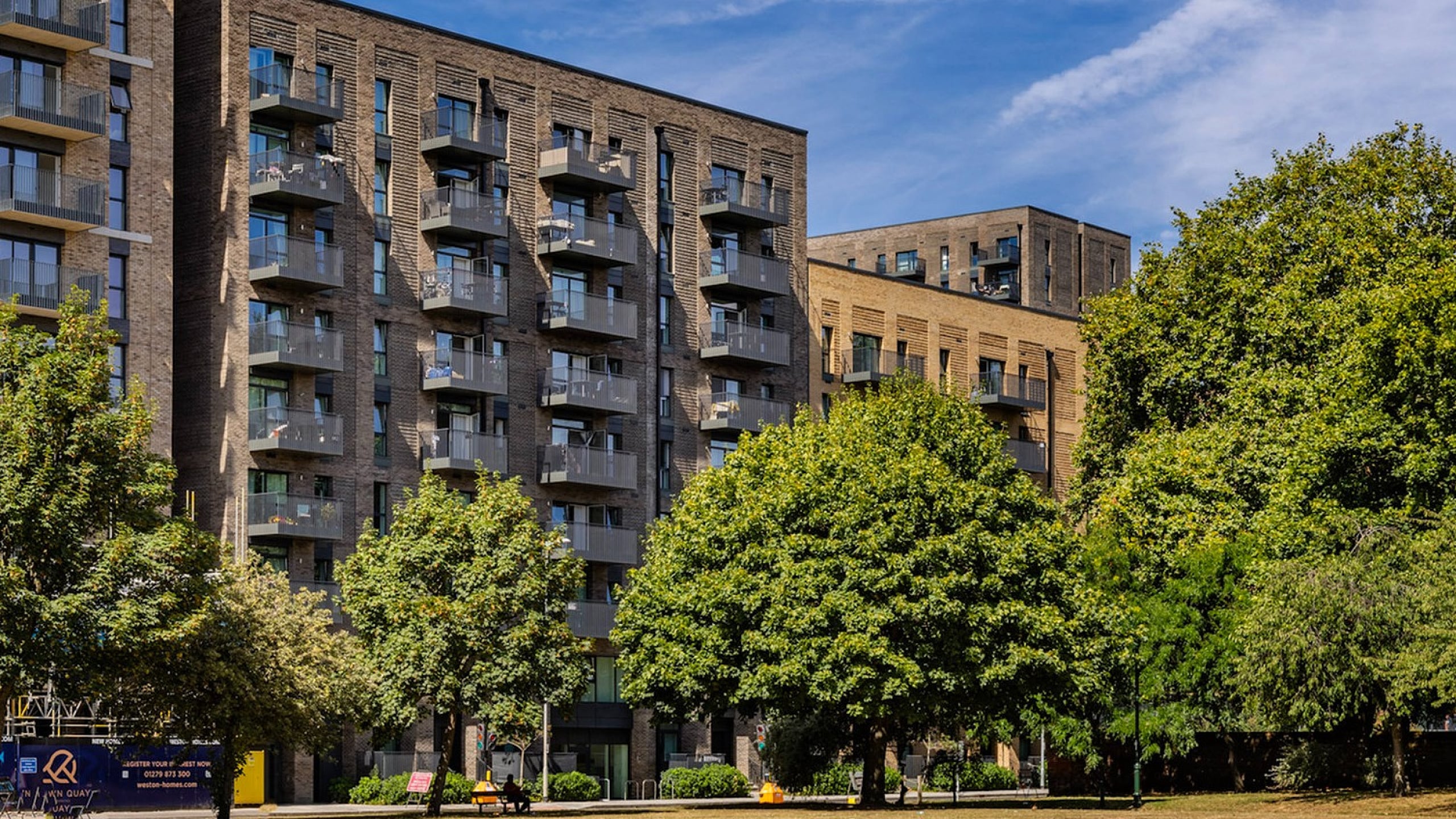 Modern urban apartment complex with stylish balconies surrounded by lush greenery and a spacious open park area under a clear blue sky.