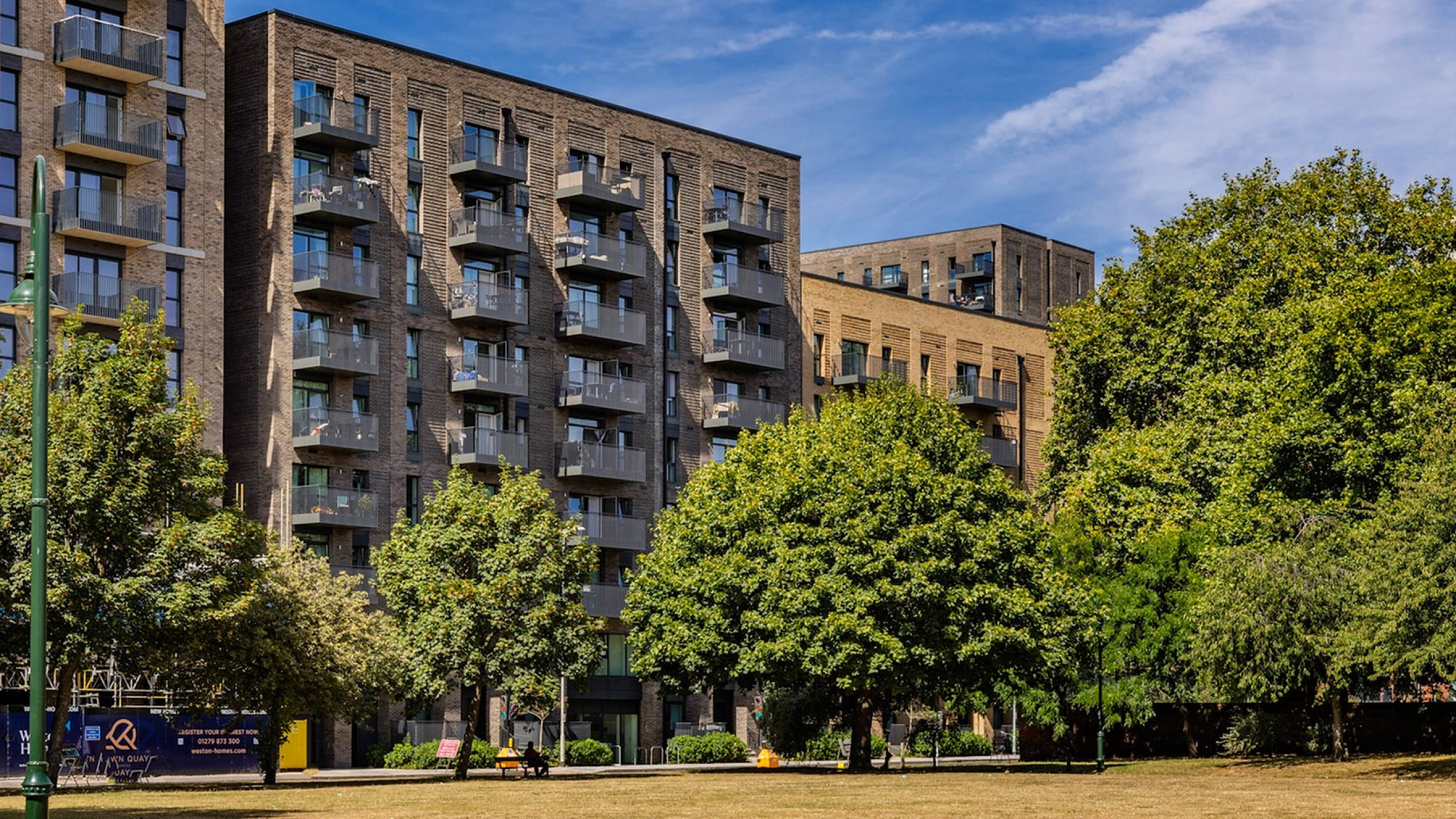 Modern apartment complex with spacious balconies, surrounded by lush greenery and a sunny park view.