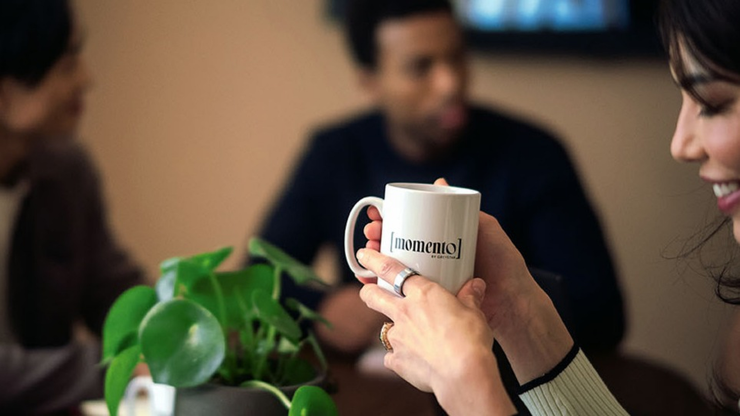 Cozy office meeting scene with smiling colleagues enjoying coffee and conversation around a table with a vibrant green plant.