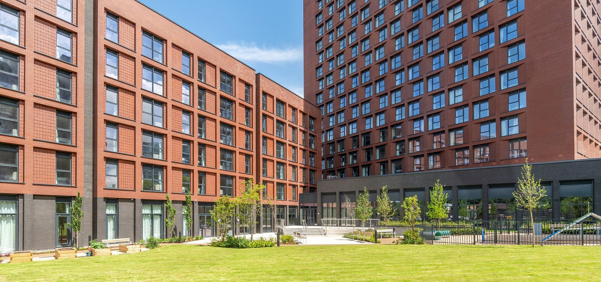 Modern apartment complex with sleek red brick facade, large windows, and beautifully landscaped green courtyard.