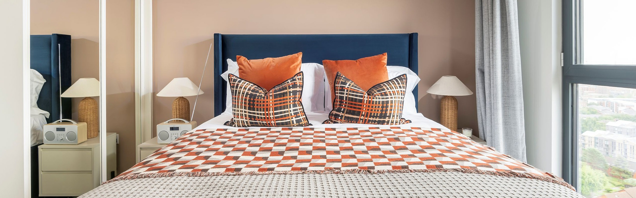 Bright and modern bedroom featuring a plush bed with vibrant cushions and a stylish patterned throw, complemented by mirrored closets and a sunlit window view.